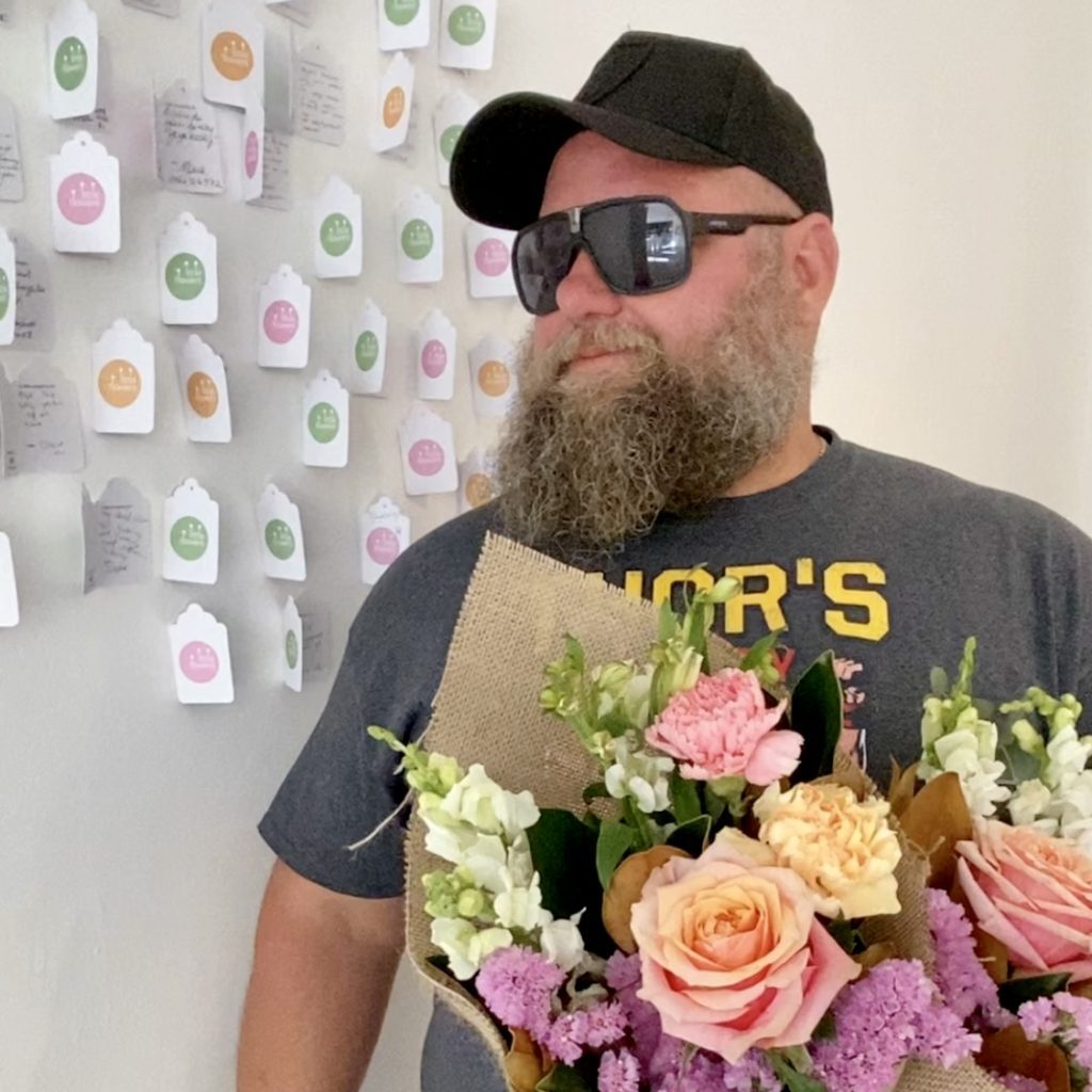 Man with beard standing in front of Gratitude Wall holding flowers