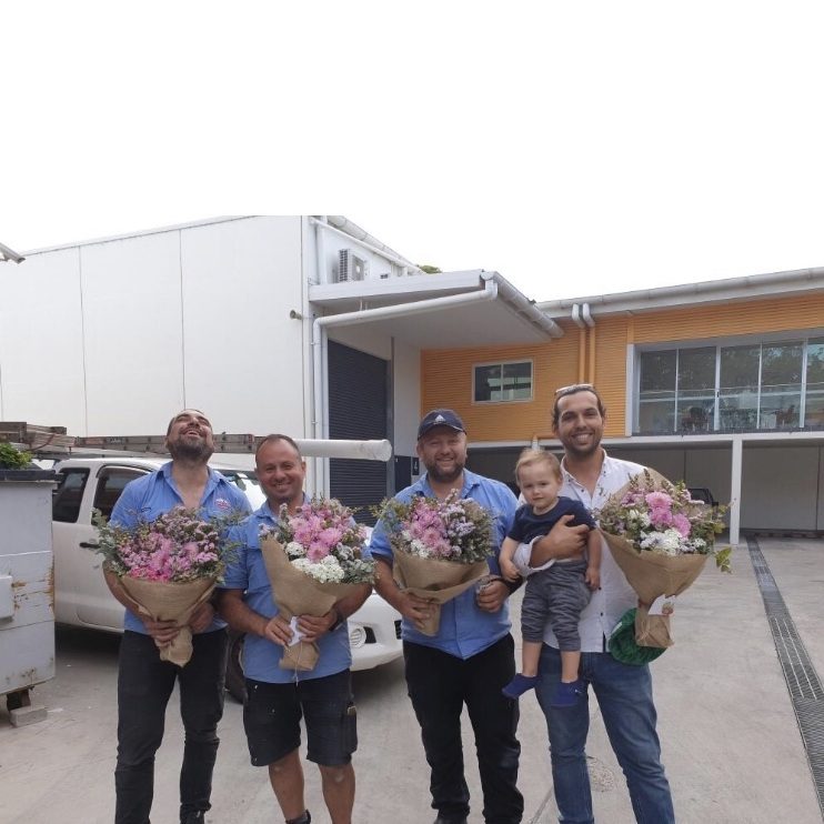 Group of men in blue work shirts smiling and holding flower bouquets, one holding a toddler.