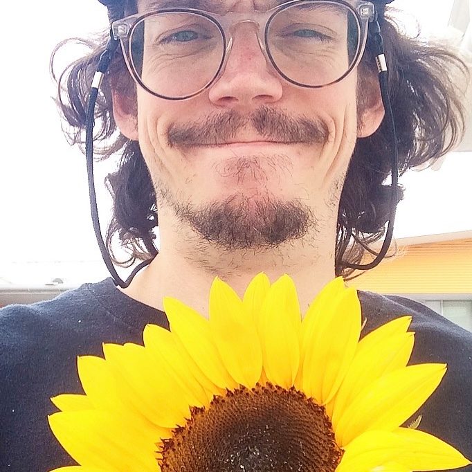 Close-up selfie of man smiling with a bright yellow sunflower in the foreground.