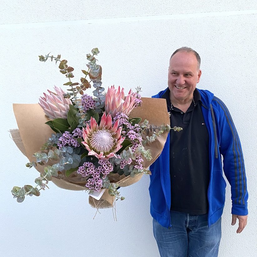 Man smiling while holding a large native flower bouquet featuring king proteas and eucalyptus.