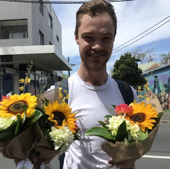 Young man smiling outdoors, holding two sunflower bouquets.